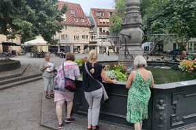 Das Bild zeigt den Brunnen auf dem Hafenmarkt. Um diesen stehen Personen und bei Ihnen steht eine Stadtführerin.
