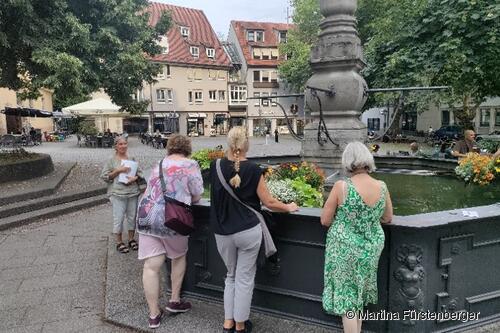 Das Bild zeigt den Brunnen auf dem Hafenmarkt. Um diesen stehen Personen und bei Ihnen steht eine Stadtführerin. Das Bild zeigt den Brunnen auf dem Hafenmarkt. Um diesen stehen Personen und bei Ihnen steht eine Stadtführerin.